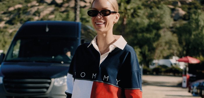 Lachend persoon met een zonnebril en een kleurblok Tommy-sweatshirt staat buiten met bomen en een bestelwagen op de achtergrond.