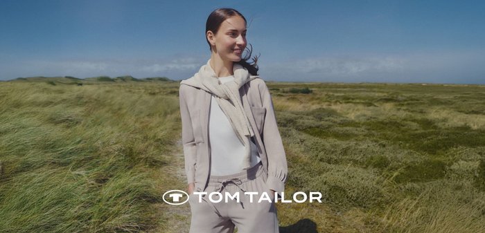 Nuori nainen seisoo nurmikkoisessa maisemassa yllään vaaleanbeige rento takki, valkoinen toppi, yhteensopivat housut ja hartioillaan huivi.