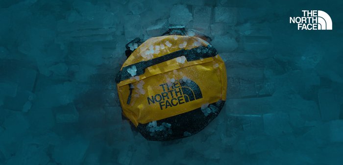 Gelbe Duffel-Tasche mit schwarzen Akzenten, ausgestattet mit dem North Face-Logo. Texturiertes Material, runde Form und detaillierte Reißverschlussbeschläge sind sichtbar.