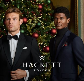 Two men in formal wear stand in front of a decorated Christmas tree. One wears a black tuxedo with a bow tie; the other, a navy suit with a turtleneck.