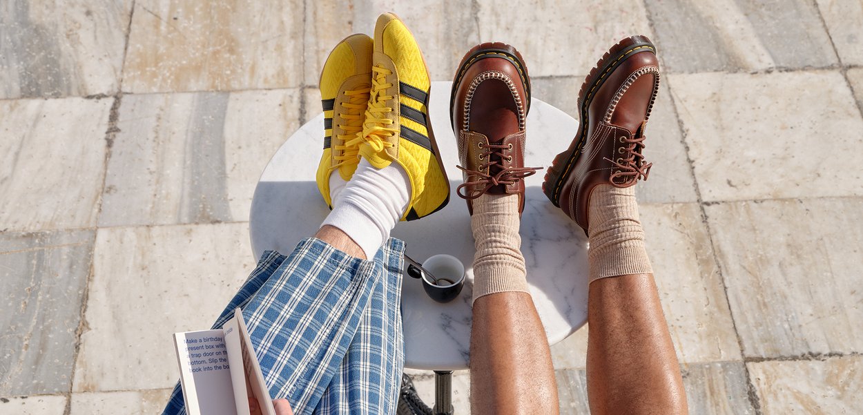 Dos personas sentadas con las piernas apoyadas sobre una mesa de mármol; una lleva zapatillas amarillas y pantalones a cuadros, la otra botas marrones y calcetines beige.