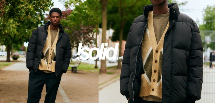 Schwarze Steppjacke über einem hellbraunen, gemusterten Strickjacke mit braunen Knöpfen. Draußen mit Bäumen und einem Weg im Hintergrund.