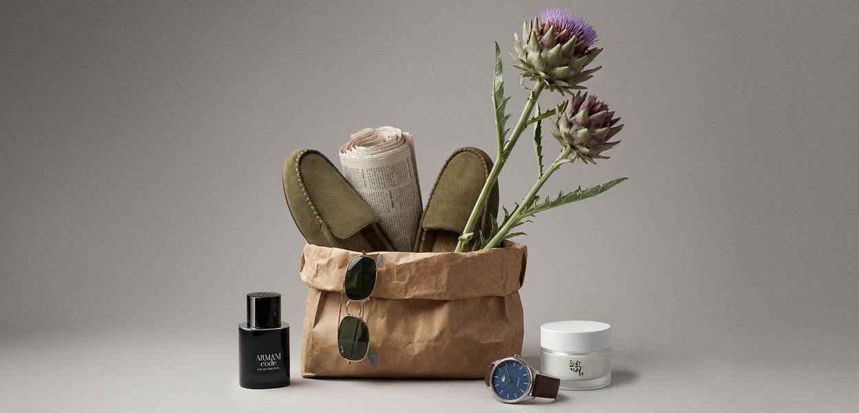 Borsa di carta marrone che contiene pantofole verdi, giornale arrotolato e fiori di cardo viola, con occhiali da sole, orologio, profumo e crema nelle vicinanze.