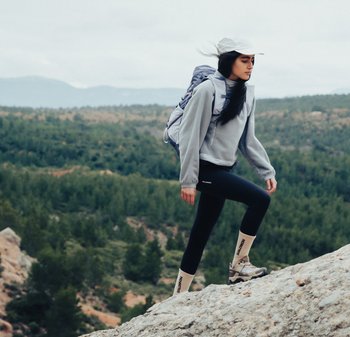 Femme portant un sac à dos et une casquette, randonnée en montée sur un terrain rocheux avec des collines boisées en arrière-plan.