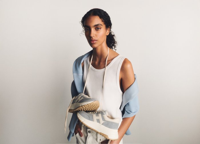 Young woman in a white vest and light blue jumper holds grey trainers hanging by their laces around her neck against a plain background.
