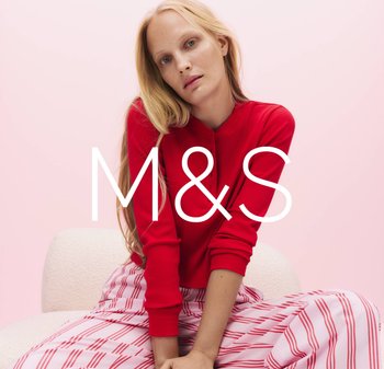 Blonde woman wearing a red long-sleeve top and pink-and-red striped pants sits on a white textured chair against a pink background.