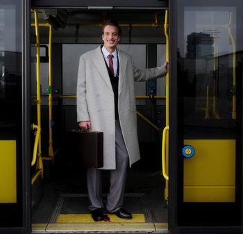 Γκρι παλτό, μαύρο γιλέκο, λευκό πουκάμισο και κόκκινος γραβάτα. Κρατώντας μια καφέ τσάντα, στέκεται στην πόρτα λεωφορείου με κίτρινες λεπτομέρειες.