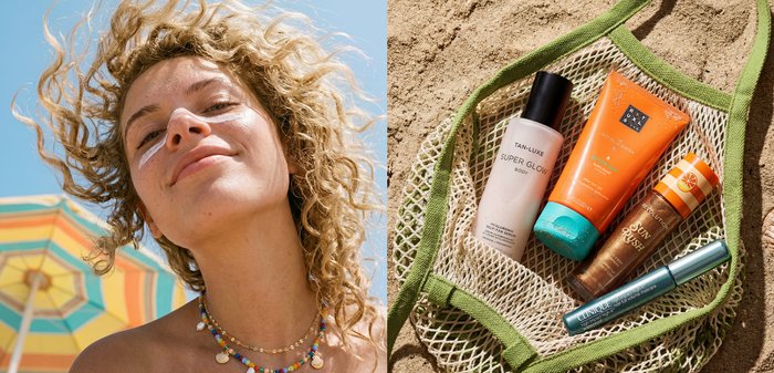Femme aux cheveux bouclés souriant en plein air près d'un parasol de plage rayé ; crèmes solaires et produits de soin de la peau disposés sur un sac en mesh vert sur le sable.