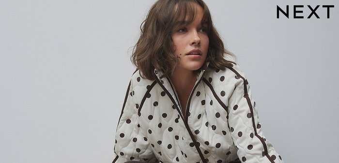 Nő ül egy barna bőrszofán, fehér, tűzött dzsekit visel fekete pöttyökkel, valamint sötét nadrágot fehér csíkokkal.