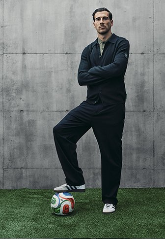 Sportowiec w ciemnej koszulce polo i szerokich spodniach stoi na trawie z kolorową piłką do soccera, która ma teksturowaną powierzchnię i wyraźne wzory.