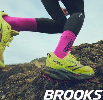 Geel hardloopschoen met zwarte accenten en een roze zool, gecombineerd met felroze sokken met het 'BROOKS' logo, gedragen op rotsachtig terrein.