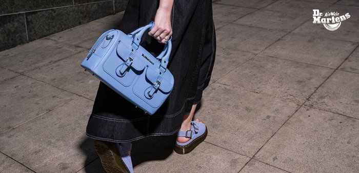 Persona caminando por la acera con sandalias azul claro y falda negra, llevando un bolso azul claro con hebillas y detalles de cremallera.