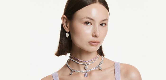 Young woman with short dark hair wears three layered gemstone necklaces and a single hoop earring against a plain background.