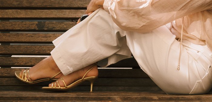 Osoba siedząca na drewnianej ławce, ubrana w białe spodnie, beżową bluzkę i złote sandały na obcasie z paskami, z jedną nogą skrzyżowaną na drugą.