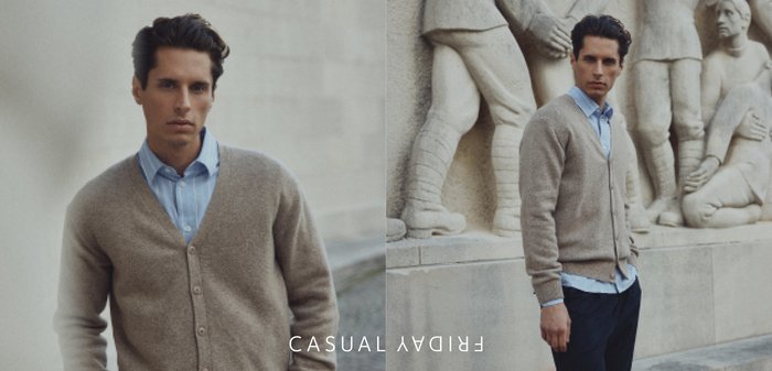 Mann trägt beige Cardigan über blauem Hemd und dunkler Hose, steht vor einer Steinwand mit Skulpturen menschlicher Figuren.