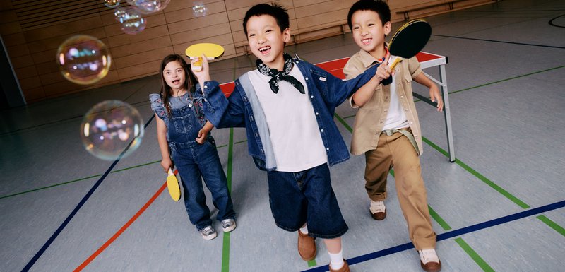Trois enfants jouent à l'intérieur, tenant des raquettes jaunes. Ils portent des vêtements en denim, une chemise khaki et un t-shirt blanc. Des bulles sont présentes.