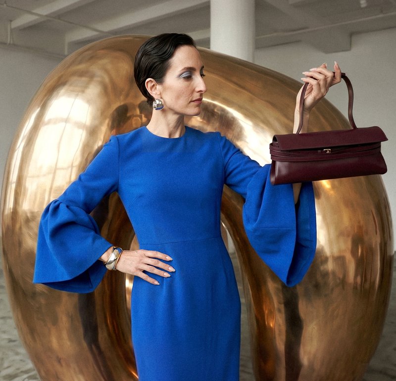 Donna con vestito blu che tiene una piccola borsa bordeaux all'altezza degli occhi, in piedi davanti a una grande scultura astratta dorata in interno.