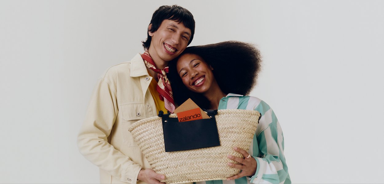 Két mosolygó ember egy nagy fonott kosarat tart, amelyben egy fekete tasak található, benne egy narancssárga Zalando borítékkal.