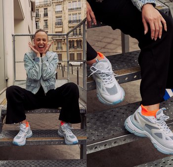 White athletic sneakers feature a mesh upper with grey accents, blue sole, and neon orange details. Equipped with laces and distinctive branding.