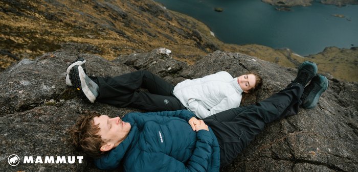 Mammut Daunenjacken in Dunkelblau und Weiß mit schwarzen Hosen, die auf felsigem Terrain nahe einem See ruhen und eine robuste Outdoor-Umgebung präsentieren.