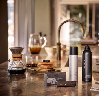 Des désodorisants d'air Rituals dans des canisters élégants de couleur grise et noire sont placés sur une surface en marbre à côté de pancakes, d'un verre et d'une cafetière.