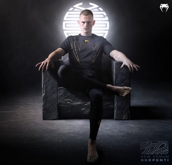 Schwarzes Sportshirt mit goldenen Akzenten und strukturiertem Design, kombiniert mit schwarzen Leggings. Modell sitzt auf einem steinartigen Stuhl vor einem dunklen Hintergrund.