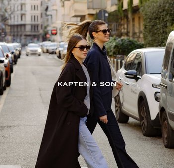 Dvě ženy v slunečních brýlích a kabátech přecházejí městskou ulici lemovanou zaparkovanými auty a budovami za denního světla.