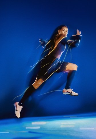 Αθλητική φιγούρα με μαύρο crop top και σορτς, φοράει λευκά παπούτσια Nike με τάπες, τρέχει μπροστά σε μπλε φόντο με δυναμικά φώτα που σχηματίζουν ιχνη.