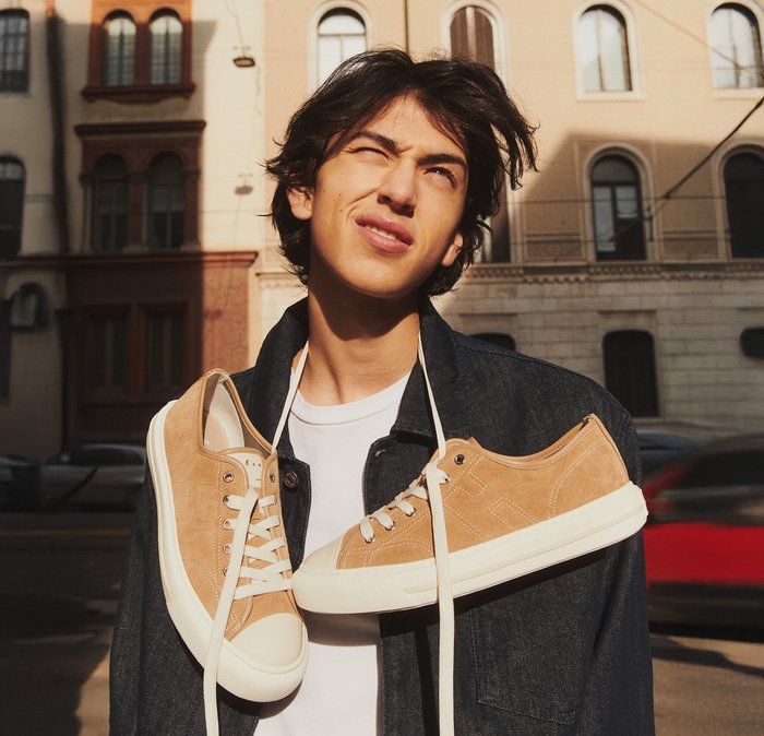 Giovane uomo all'aperto che strizza gli occhi alla luce del sole con scarpe da ginnastica color sabbia appese al collo tramite i lacci, sullo sfondo di un edificio cittadino.