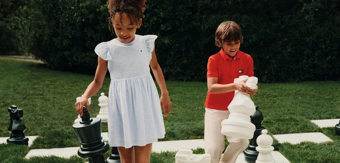 Zwei Kinder im Freien, die große Schachfiguren auf einem lebensgroßen Schachbrett mit Gras- und Heckenhintergrund halten.