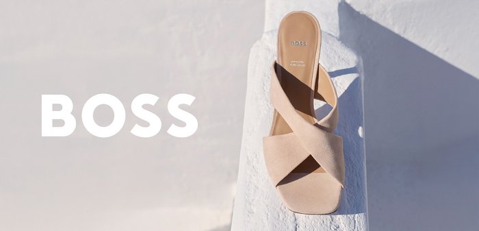 Beige suède BOSS-sandaal met gekruiste bandjes die op een witte getextureerde ondergrond ligt, vet wit BOSS-logo links op een lichte achtergrond.