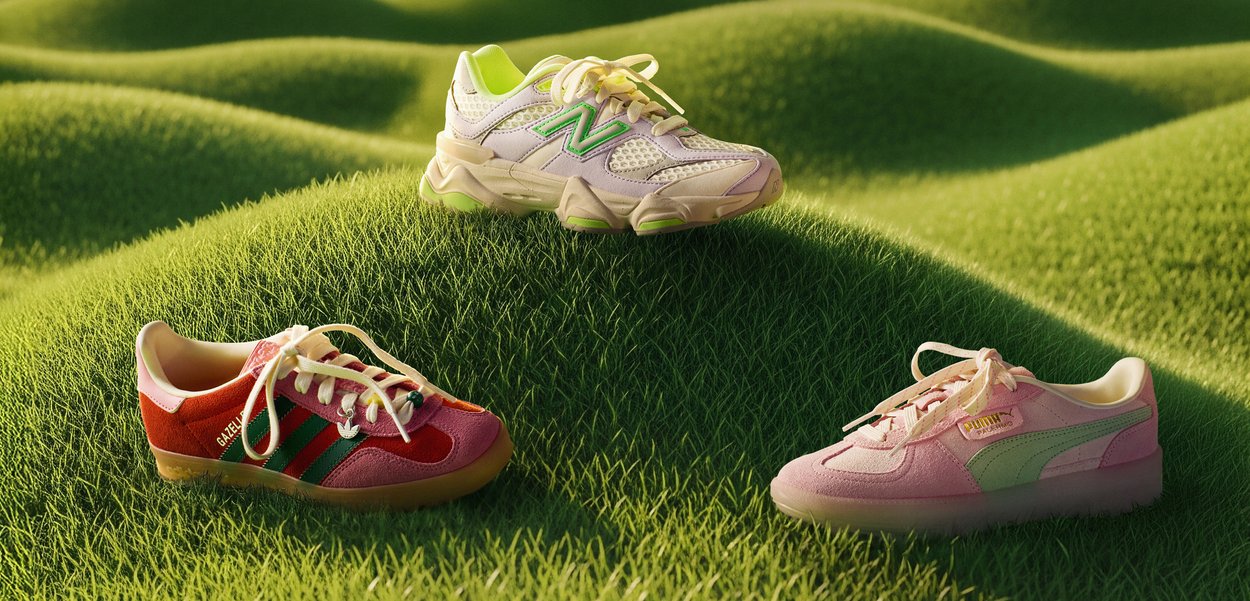 Drie sneakers op groen gras: een New Balance-model in wit met groene accenten, een Adidas Gazelle in rood en roze, en een roze Puma-stijl met mintaccenten.