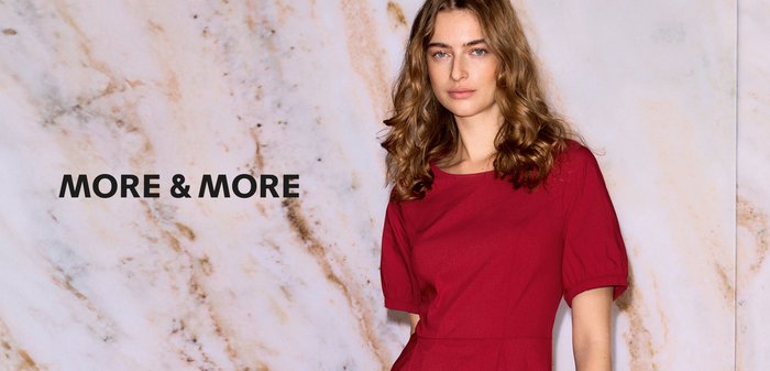 Junge Frau mit welligem Haar steht in einem roten knielangen Kleid vor einem hellen Marmohintergrund mit dem Text "MORE & MORE".