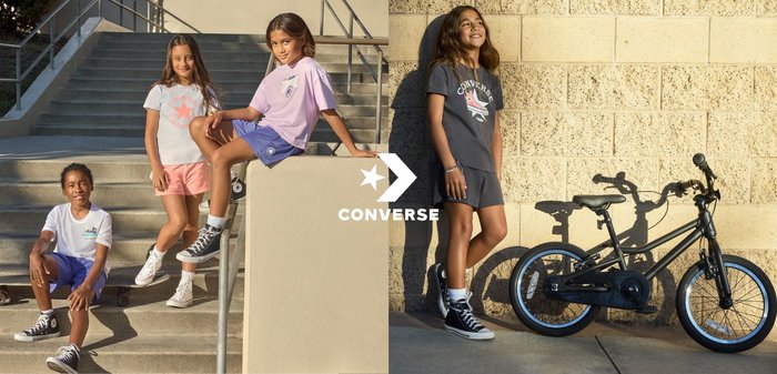 Jeune personne aux cheveux blonds effectue un trick de skateboard en l'air devant un mur en briques claires, portant des chaussures et des vêtements Converse noirs.