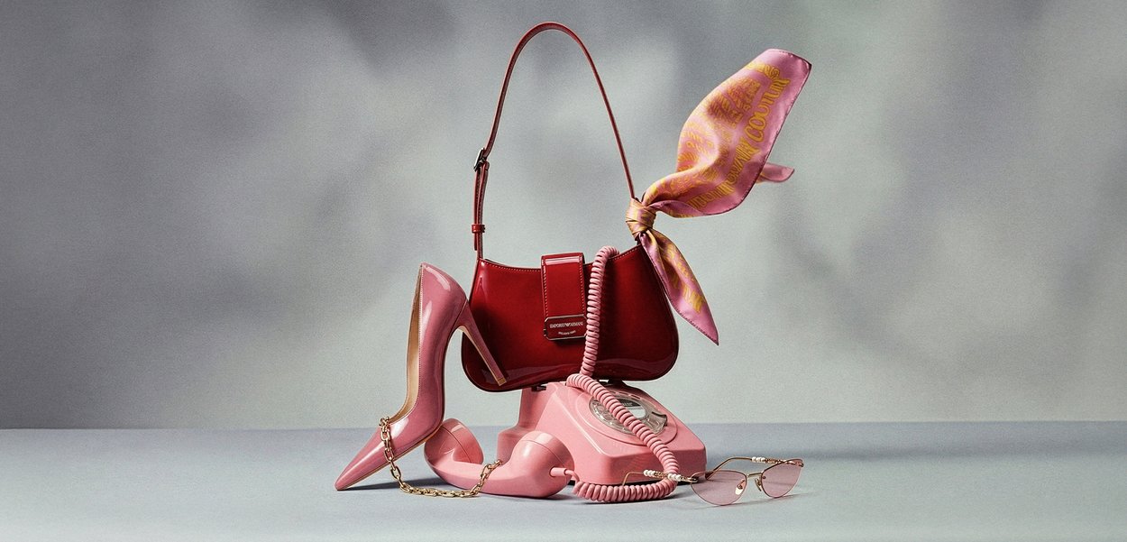 Błyszcząca czerwona torebka z różowym szalikiem zawiązanym na uchwycie, umieszczona na różowym, retro telefonie obrotowym, obok różowego szpilka i różowych okularów przeciwsłonecznych z różowymi szkłami.