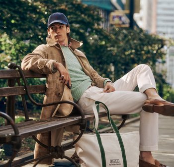 Joven con un abrigo marrón, suéter verde, pantalones blancos y gorra sentado en un banco con un gran bolso Polo a su lado.