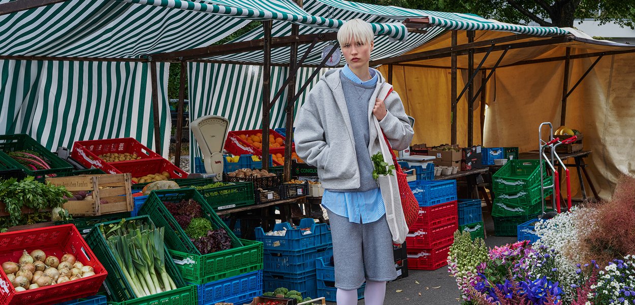 Hanorac gri cu glugă și pantaloni scurți peste o cămașă albastră deschisă, ținând o pungă roșie din plasă cu ierburi la un market vibrant cu un cort cu dungi.