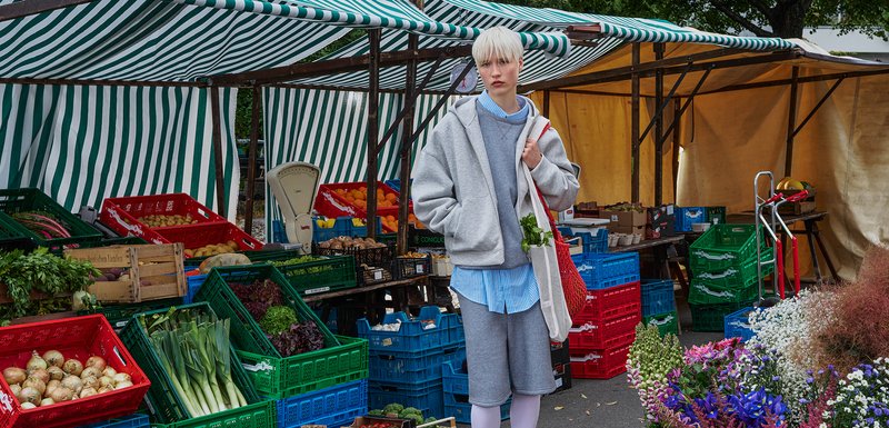 Sweat à capuche gris et shorts sur une chemise bleu clair, tenant un sac en maille rouge rempli d'herbes dans un marché animé sous un auvent rayé.