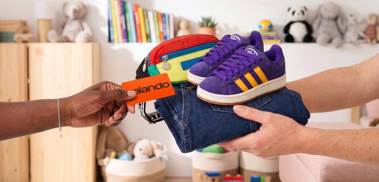 Due mani si scambiano jeans piegati, scarpe da ginnastica viola, una borsa colorata e una carta Galando arancione in una stanza dei bambini con giocattoli e libri.