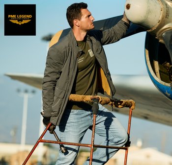 Man in een casual jas en spijkerbroek controleert het vliegtuigmotor terwijl hij op een roestige onderhoudsladder buiten in daglicht staat.