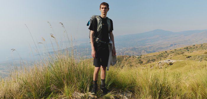 Young man standing on a grassy hillside wearing hiking gear and carrying a backpack with mountains visible in the distance.