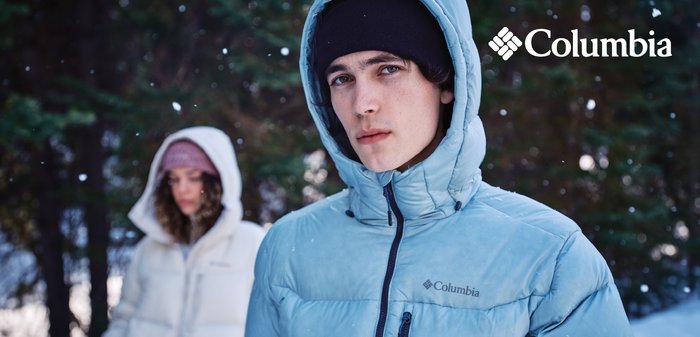 Zwei Personen im Freien in einem verschneiten Wald, die Columbia Winterjacken und Mützen tragen, während leise Schnee um sie herum fällt.