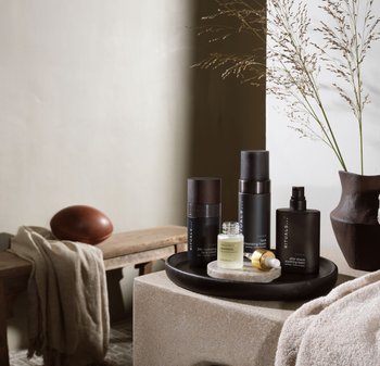 Sort bakke med fire hudplejeprodukter: to mørke flasker med træpropper, en lille glaskrukke og en tube. Beige stenshoverflade og blødt klæde.