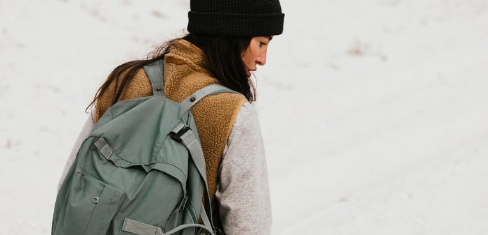 Grüner Rucksack mit mehreren Taschen, getragen über einem braunen Fleece-Weste und einem grauen Langarmshirt, vor einem schneebedeckten Hintergrund.