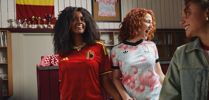 Trois femmes souriantes se tenant la main à l'intérieur, deux portant des maillots de football de Belgique et d'Argentine, avec des trophées et un drapeau en arrière-plan.