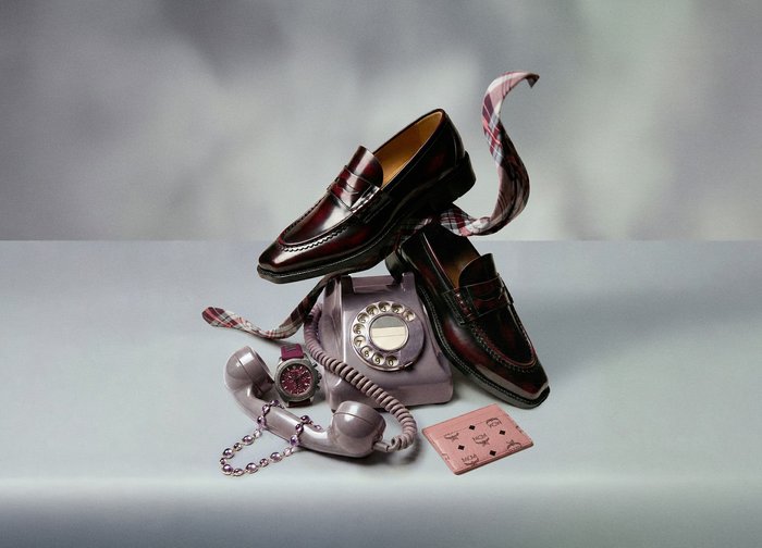 Paire de mocassins en cuir bordeaux posée sur un téléphone vintage à cadran rotatif, accompagnée d'une montre assortie, d'une écharpe à carreaux et d'un porte-cartes rose sur une surface grise.