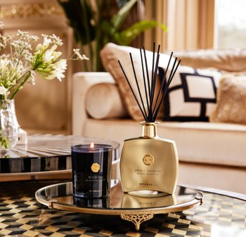 Diffuseur en verre doré avec des bâtonnets noirs étiqueté "Jasmin Doux" à côté d'une bougie bleu foncé étiquetée "Figue Sauvage" sur un plateau en miroir.