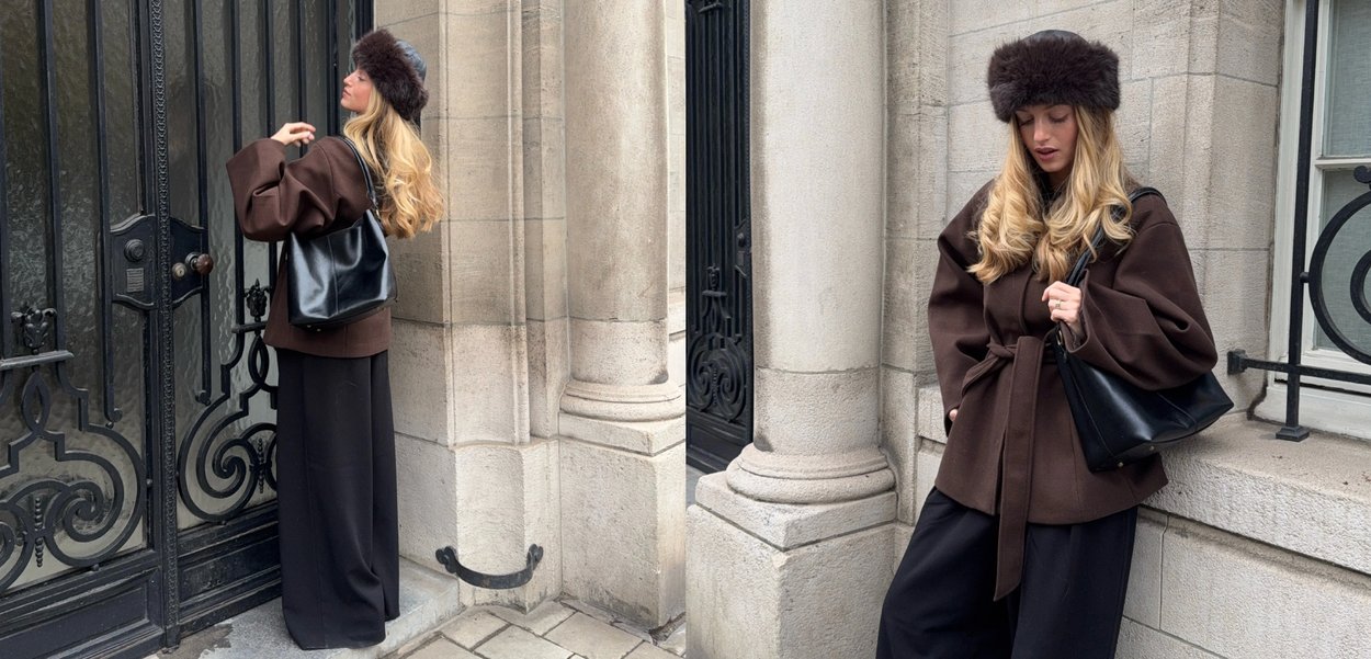 Kvinde iført brun frakke, sorte vidde bukser, pelslue og sort skuldertaske, stående ved udsmykkede sorte jernporte og stensøjler.