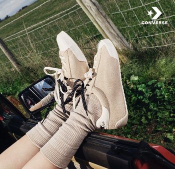 Pies con zapatillas Converse beige con cordones desparejados apoyados fuera de la ventana de un coche en una zona rural verde.