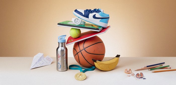 Μπάσκετ, αθλητικό παπούτσι, τετράδια, μήλο, ρολόι, μπουκάλι νερό, μετάλλιο, χάρτινο αεροπλανάκι, μπανάνα και έγχρωμα μολύβια τοποθετημένα πάνω σε ένα τραπέζι.